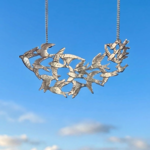 Handmade sterling silver statement pendant featuring a murmuration flock of seventeen hand-engraved birds in flight against a blue sky background