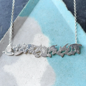 Flat lay of a handmade sterling silver starling flock bar necklace resting on a textured blue and grey ceramic dish, crafted by Stacy Bias Jewellery in Scotland.