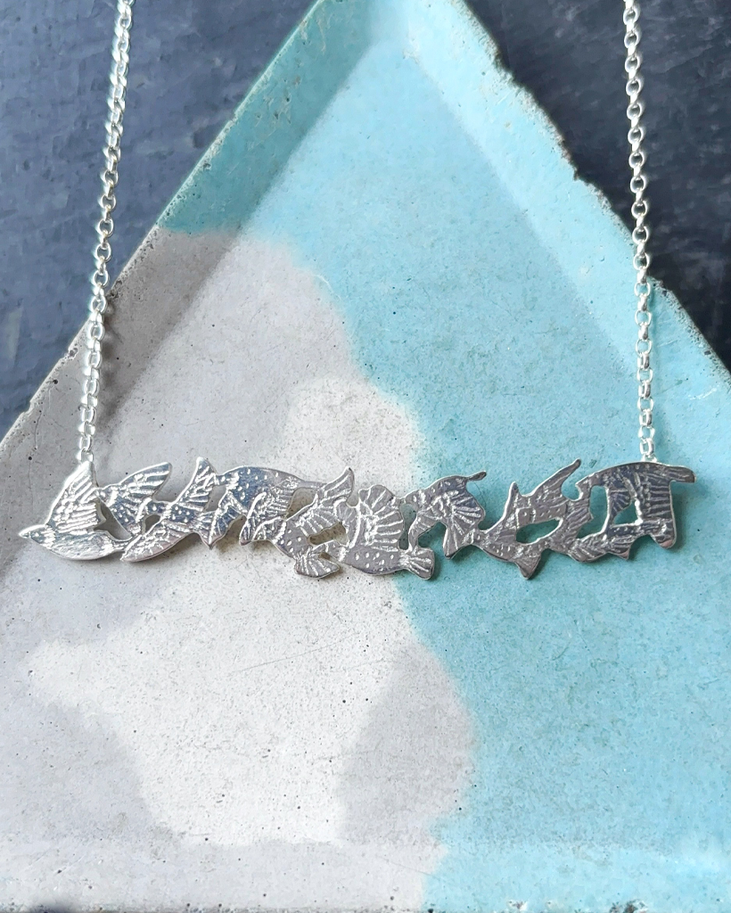 Contemporary Silver Starling Flock Necklace | Handmade Scottish Nature Jewellery Flat lay of a handmade sterling silver starling flock bar necklace resting on a textured blue and grey ceramic dish, crafted by Stacy Bias Jewellery in Scotland.