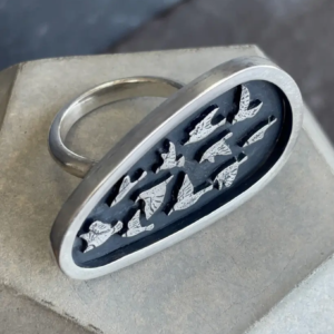 High angle shot of the sterling silver Murmuration ring showing the band construction and the oxidized shield with flying birds resting on a concrete plinth.