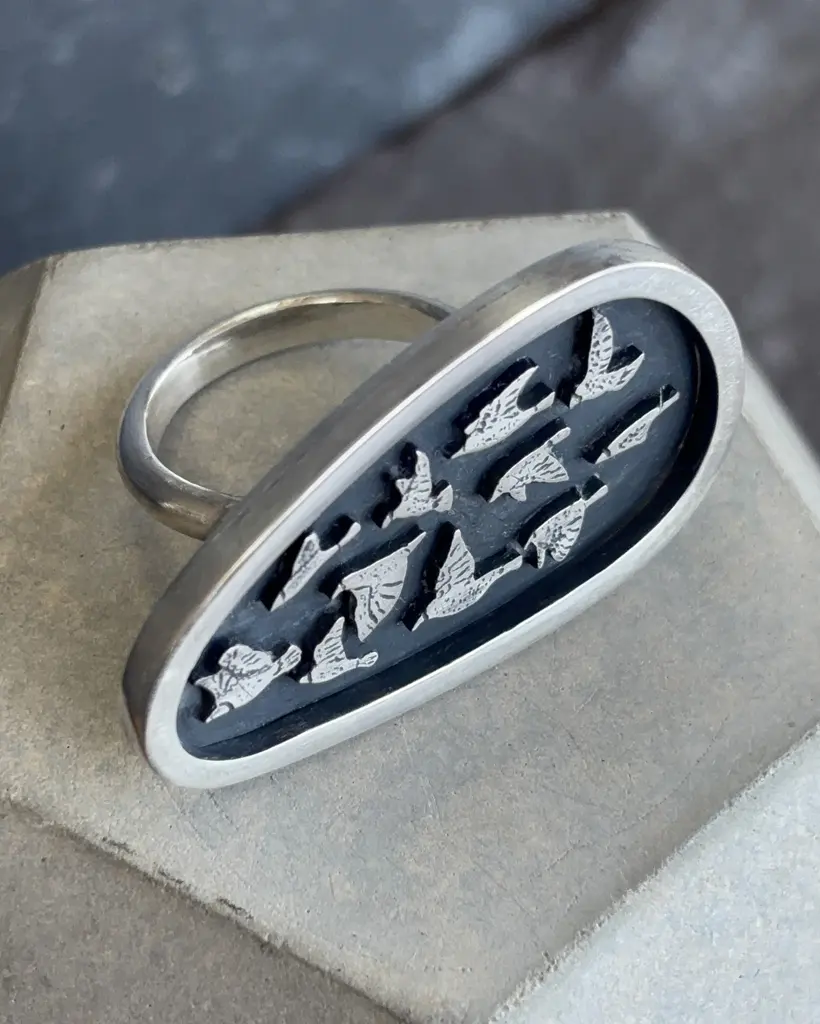 High angle shot of the sterling silver Murmuration ring showing the band construction and the oxidized shield with flying birds resting on a concrete plinth.
