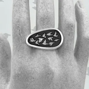 Artisan handmade sterling silver statement ring featuring an oval shield with a flock of silver birds against an oxidized black background, displayed on a mannequin hand.