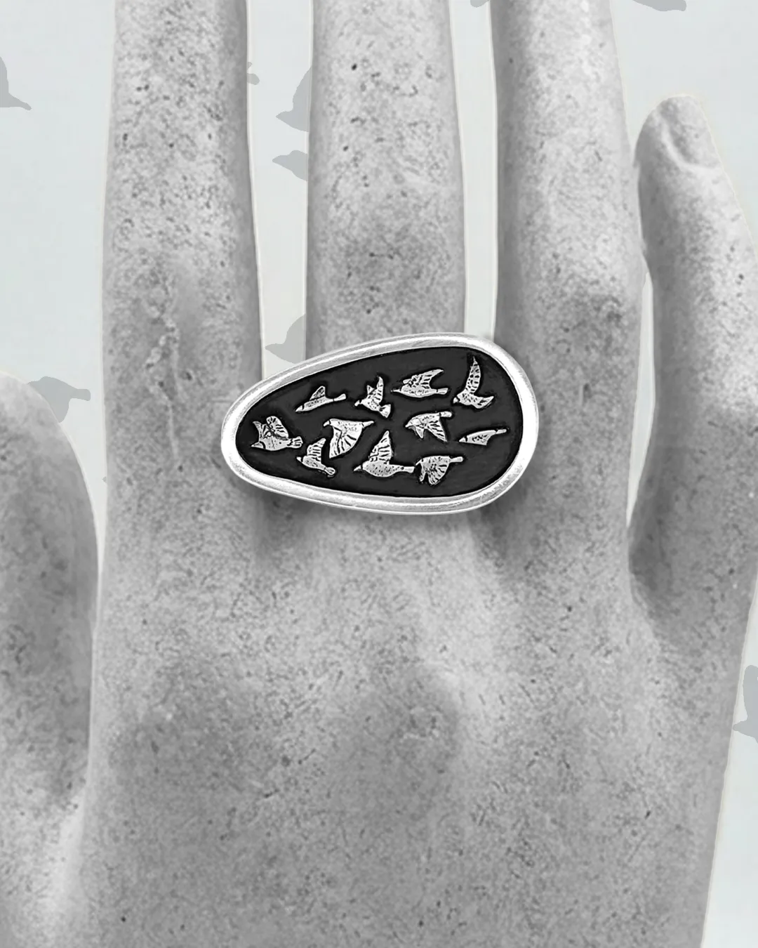 Artisan handmade sterling silver statement ring featuring an oval shield with a flock of silver birds against an oxidized black background, displayed on a mannequin hand.