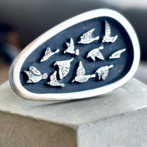 Close-up studio shot of the handmade sterling silver Murmuration ring with a textured bird flock on a dark oxidized shield, resting on a grey concrete block.