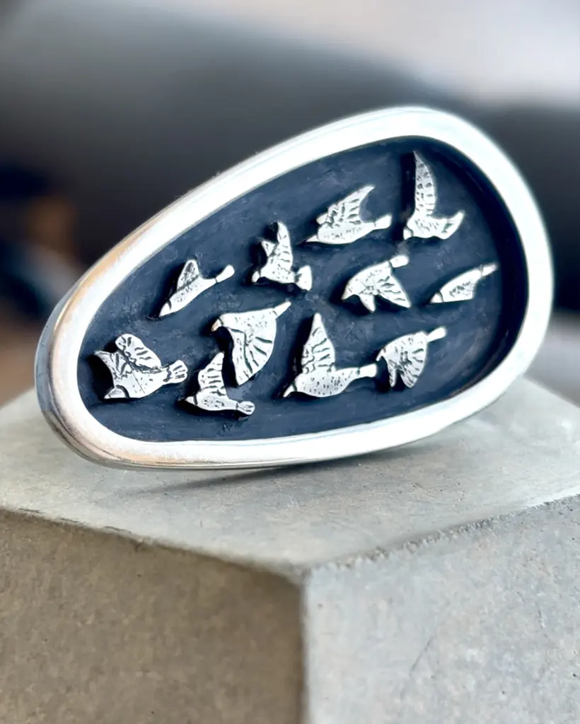 Close-up studio shot of the handmade sterling silver Murmuration ring with a textured bird flock on a dark oxidized shield, resting on a grey concrete block.