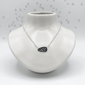 Artisan handmade sterling silver asymmetrical necklace showing a flock of birds on a black shield, displayed on a white ceramic neck bust with illustrated starlings in the background.