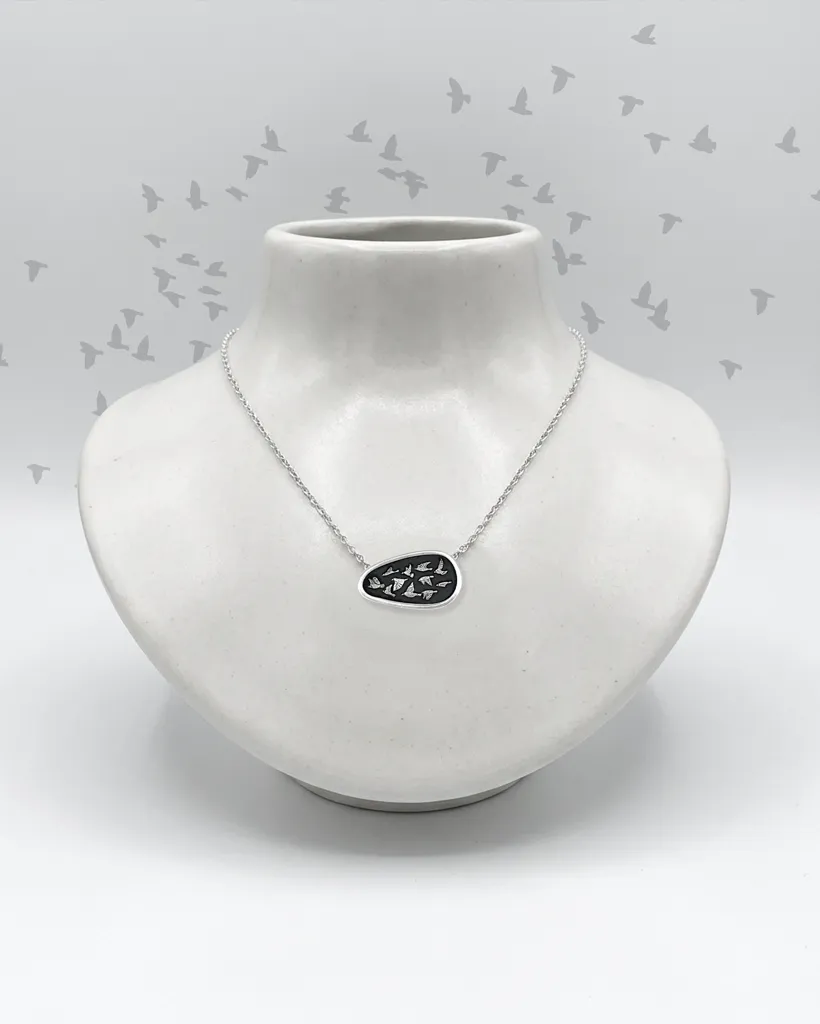 Artisan handmade sterling silver asymmetrical necklace showing a flock of birds on a black shield, displayed on a white ceramic neck bust with illustrated starlings in the background.