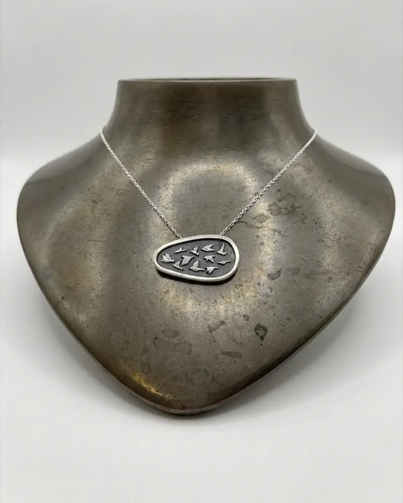 Artisan handmade sterling silver asymmetrical necklace with a flock of silver birds on an oxidized shield background, displayed on a dark metallic neck bust.