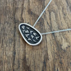 Flat lay of a handmade sterling silver asymmetrical bird shield necklace with oxidized detailing, resting on a textured rustic wooden tabletop.