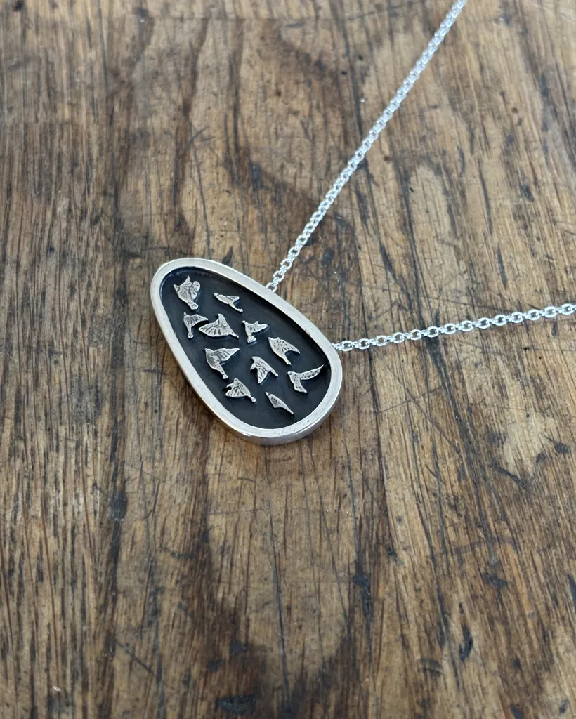 Organic Silver Murmuration Pendant | Scottish Handcrafted Bird Necklace Flat lay of a handmade sterling silver asymmetrical bird shield necklace with oxidized detailing, resting on a textured rustic wooden tabletop.