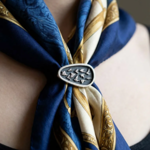 Artisan handmade sterling silver oval brooch featuring a flock of hand-engraved flying birds, pinned onto a patterned navy and gold silk scarf.