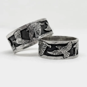 Two wide oxidised sterling silver starling band rings at a low angle, the flock of birds tracking around each band