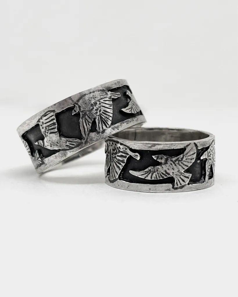 Two wide oxidised sterling silver starling band rings at a low angle, the flock of birds tracking around each band