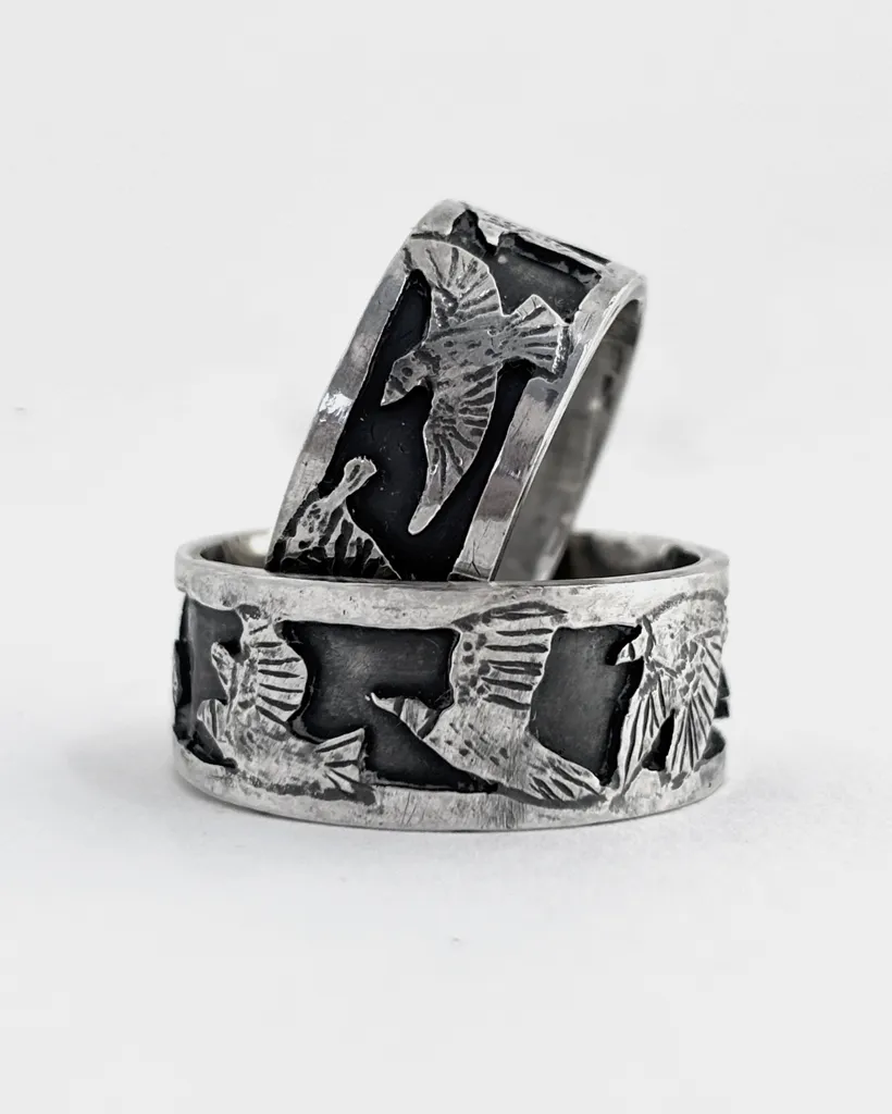 Two wide oxidised sterling silver starling band rings on white, bright raised birds in flight against a dark ground