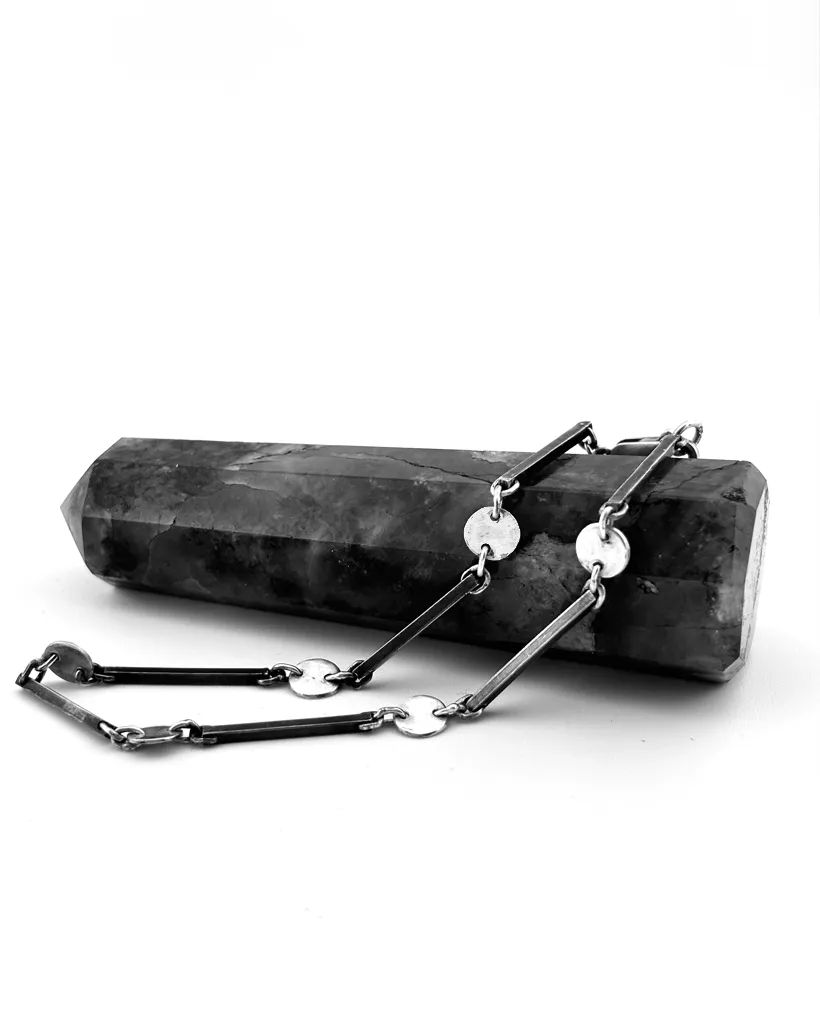 Handmade sterling silver bracelet featuring alternating thin metal bars and flat circular disc links draped over a dark crystal point.