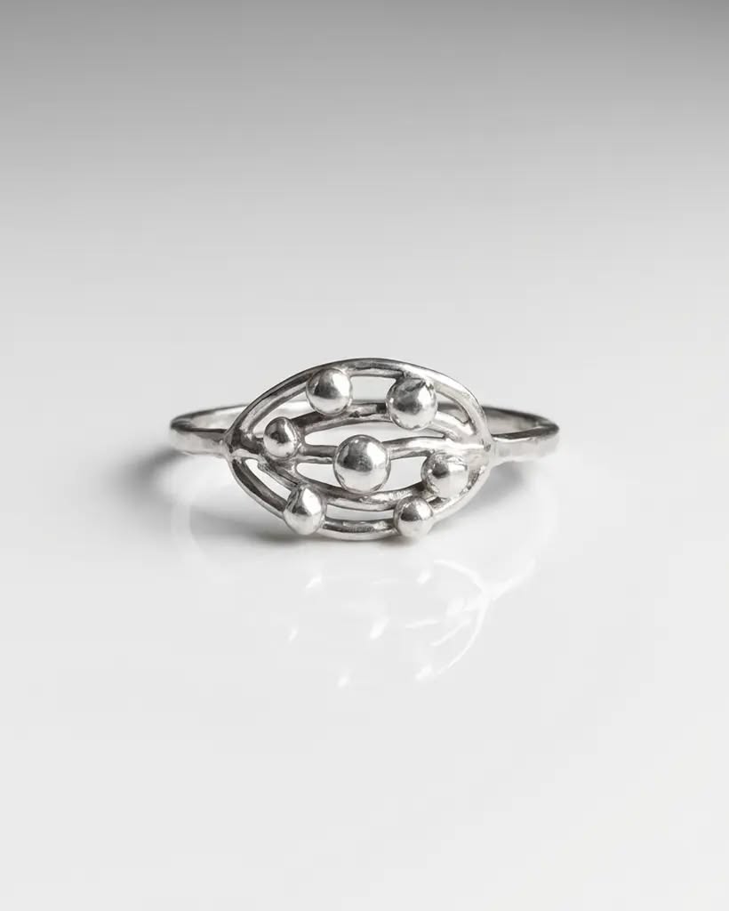 Orbit Cluster Ring in Sterling Silver | Artisan Celestial Stacking Band Close-up studio shot of the handmade sterling silver Orbit Cluster ring with reflective silver spheres inside an oval frame, resting on a white reflective surface.
