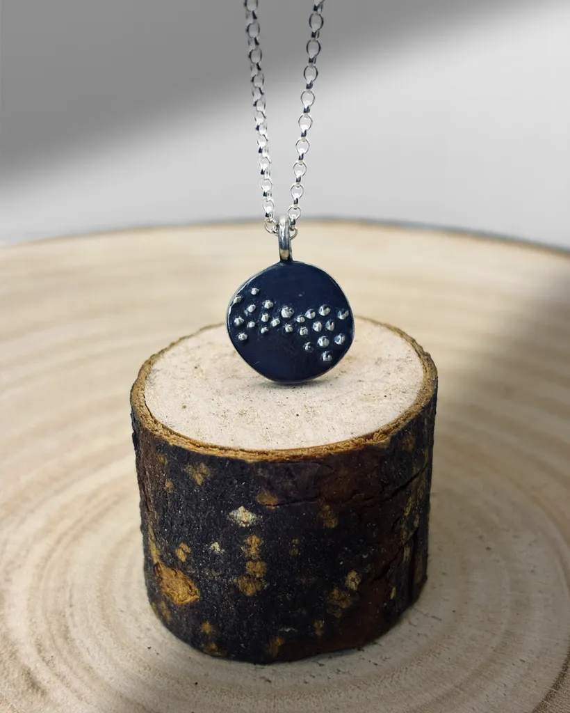 Silver Murmuration Dot Flock pendant hanging over a small wooden log display. Handcrafted by Stacy Bias.