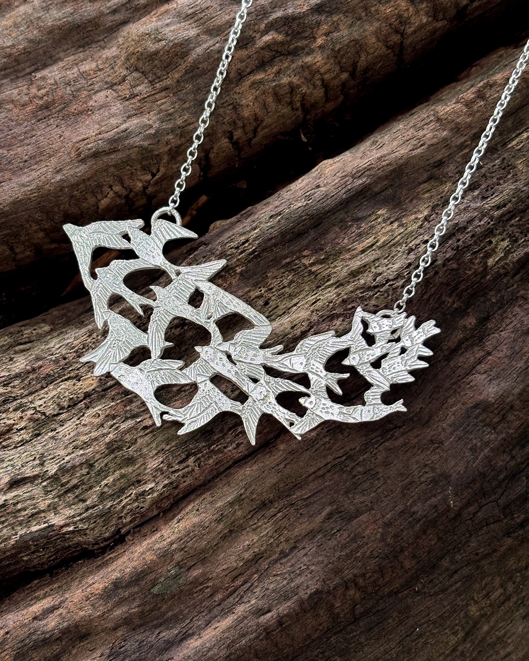 Angled closeup of a hand-pierced sterling silver statement necklace featuring a flock of starlings on a rustic wood log.