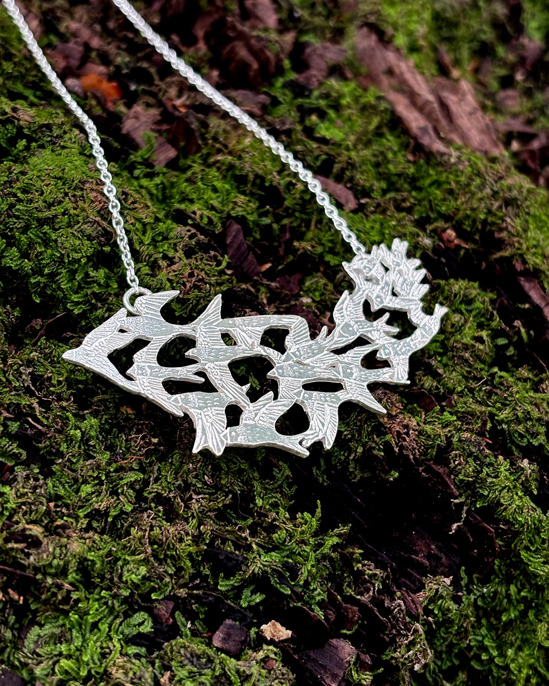 Diagonal view of the Scottish artisan silver murmuration necklace resting on moss-covered bark.
